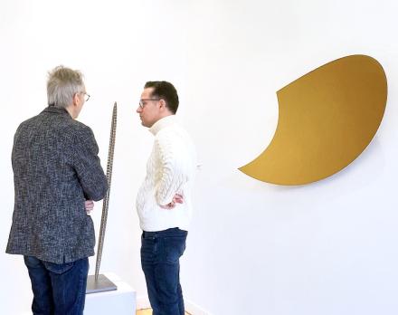 Martin Willing, Gestrecktes Ellipsoid, 1991/1999, Niro Federband, gelasert, gebogen, geschweißt, eingearbeitet in Edelstahlplatte, Höhe 127 cm, und Heiner Thiel, WVZ 795, 2025, eloxiertes Aluminium, 60 x 104 x 19 cm, mit Ausstellungsgästen